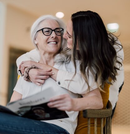 Ältere Frau mit Brille umarmt von junger Frau mit Zeitschrift im Arm | © Spa Resort Geinberg / Chris Perkles